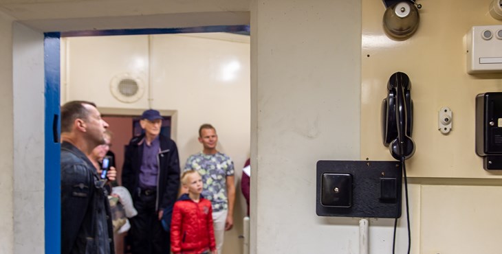 Luisterende deelnemers van de rondleiding door de ondergrondse bunker, met op de voorgrond een oude telefoon en alarmbel.