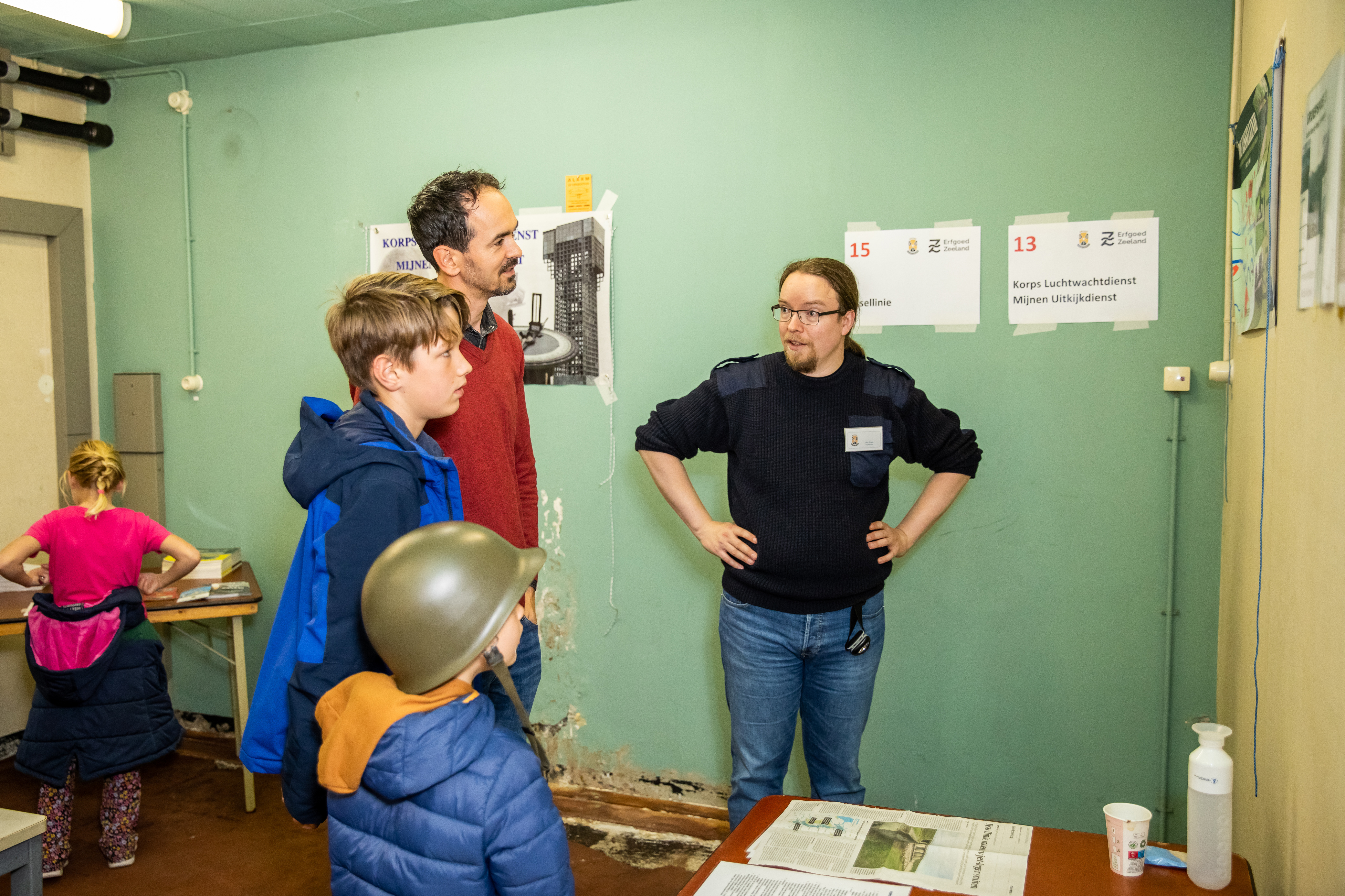 Een man en twee kinderen, een met helm op, in BB-bunker luisterend naar uitleg van vrijwilliger.