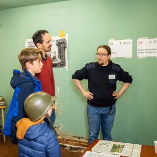 Een man en twee kinderen, een met helm op, in BB-bunker luisterend naar uitleg van vrijwilliger.