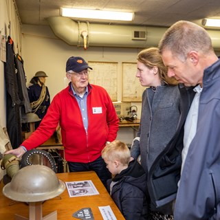 Vrijwilliger geeft uitleg aan een man, een vrouw en een jongetje over dienstspullen in BB-bunker.
