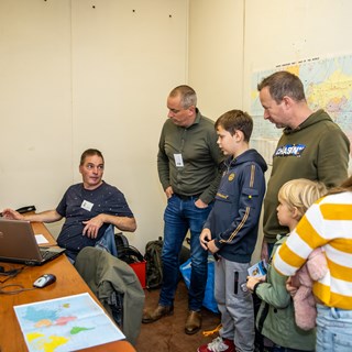 Twee mannen en drie kinderen luisteren naar uitleg van vrijwilliger in ruimte met plattegronden.