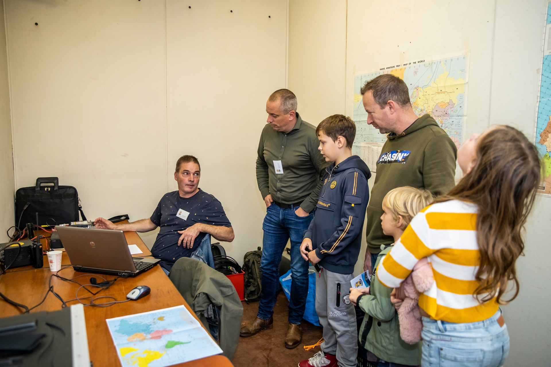 Twee mannen en drie kinderen luisteren naar uitleg van vrijwilliger in ruimte met plattegronden.
