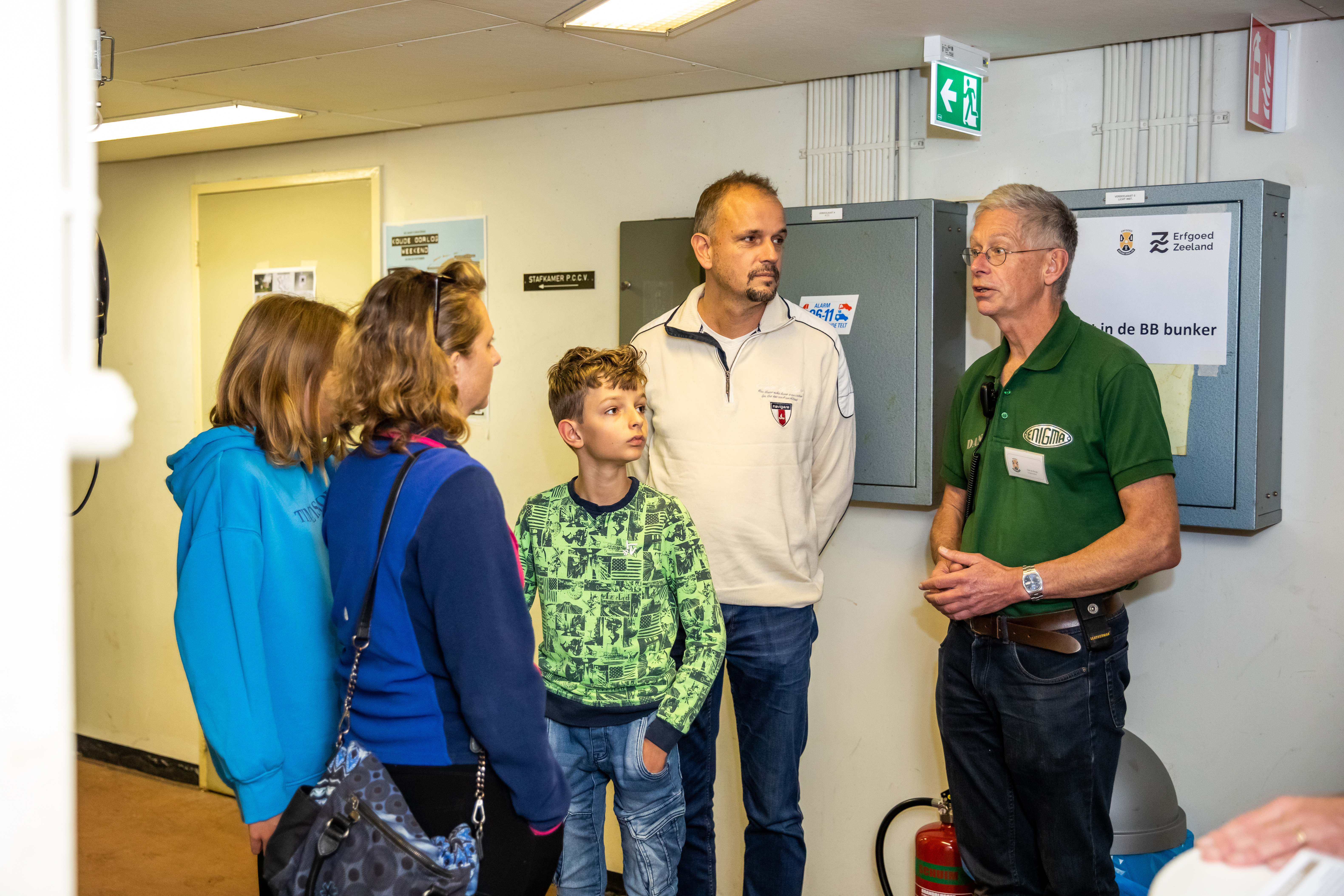 Een man, een vrouw en twee kinderen luisterend staand naar een vrijwilliger in de BB-bunker.