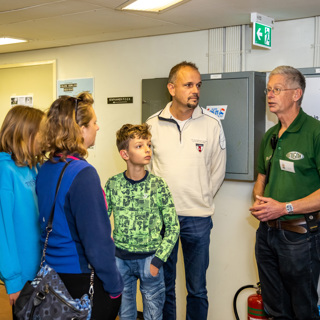 Een man, een vrouw en twee kinderen luisterend staand naar een vrijwilliger in de BB-bunker.
