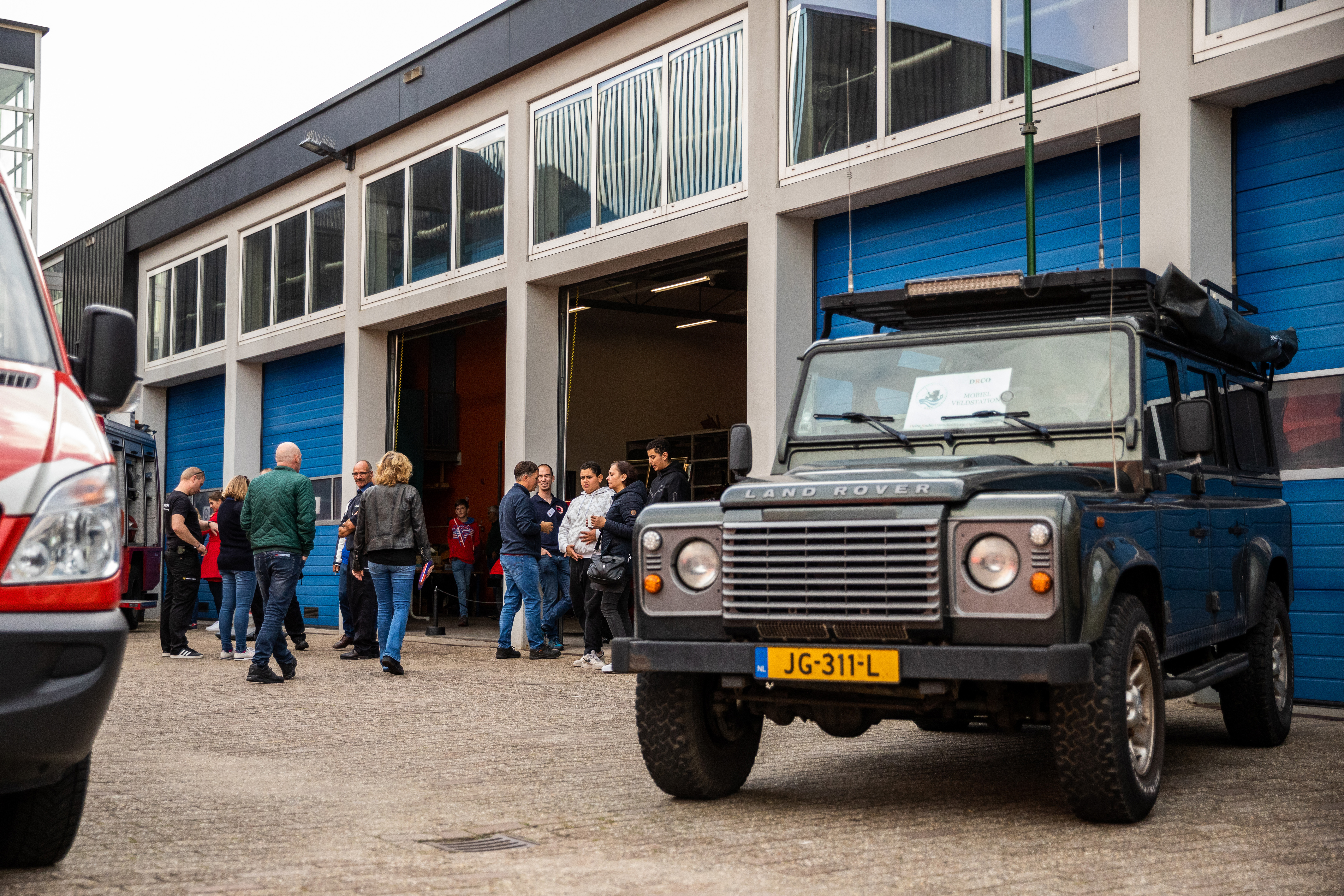 Landrover geparkeerd op het terrein voor de stallingsruimte met op de achtergrond lopende mensen.