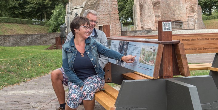 Een man en een vrouw zitten op de klapbank in Hulst en lezen over de historie, die vermeld staat op de klapbank.