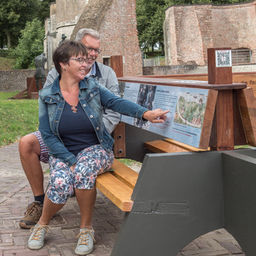 Een man en een vrouw zitten op de klapbank in Hulst en lezen over de historie, die vermeld staat op de klapbank.