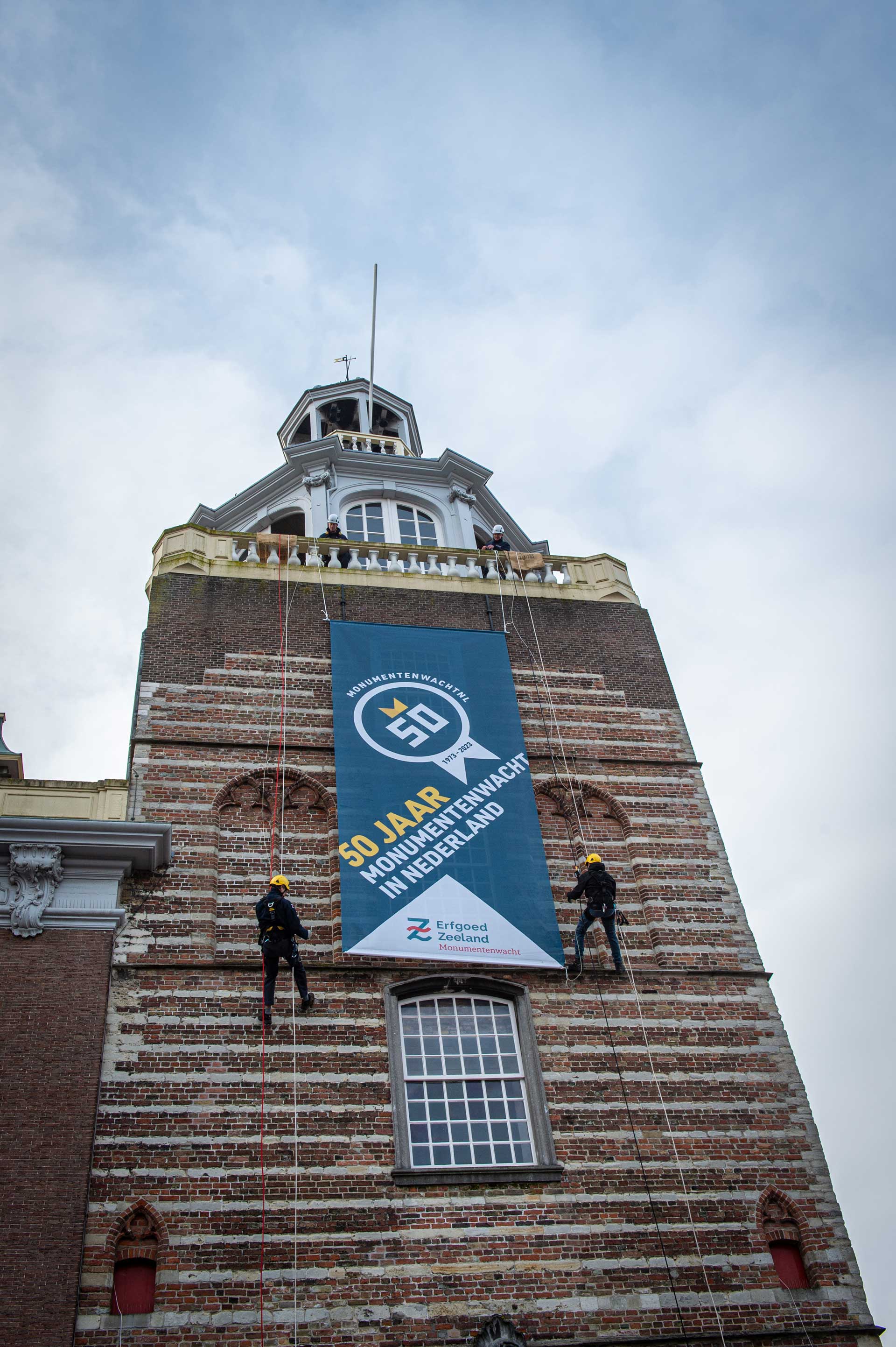 Monumentenwachters rollen de feestelijke banner '50 jaar Monumentenwacht in Nederland 1973-2023' uit aan de gevel van het stadhuis in Goes.