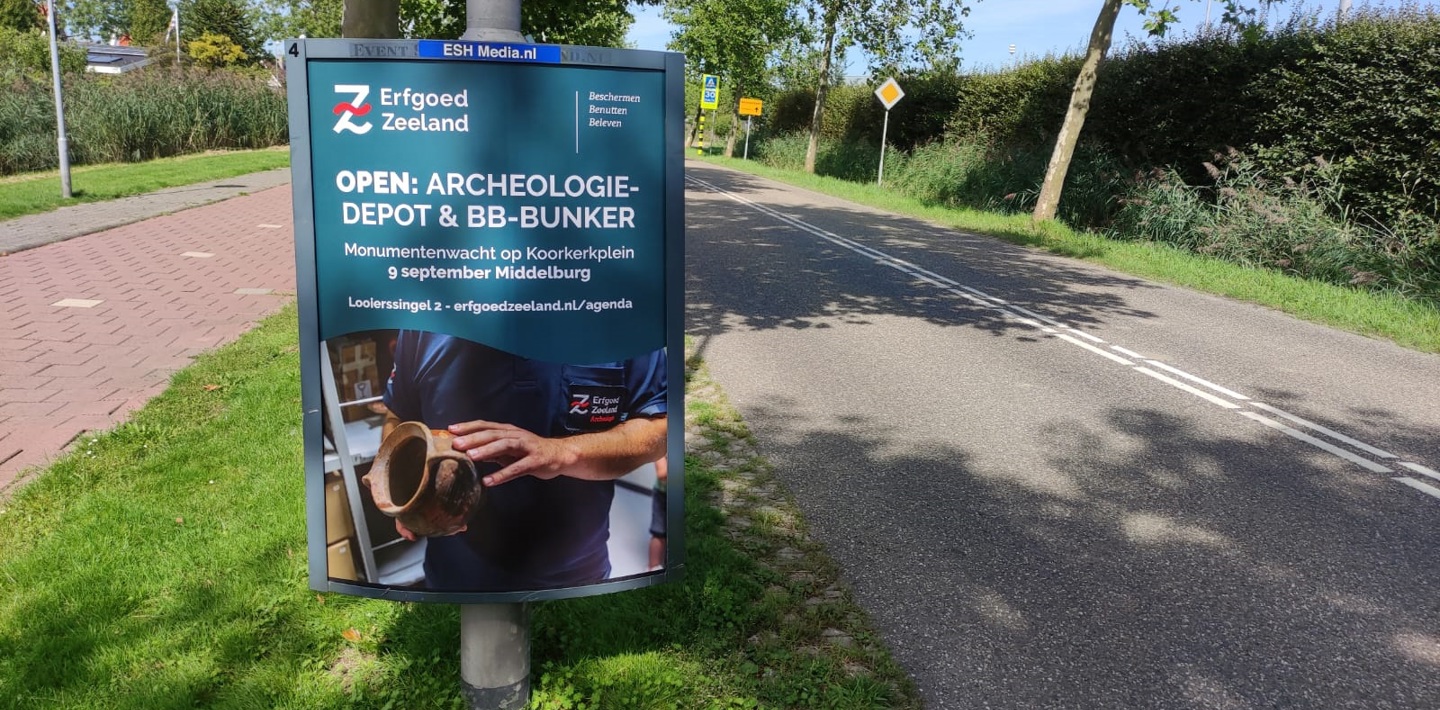 Op een reclamebord langs de weg is de poster met activiteiten te zien die Erfgoed Zeeland organiseerde tijdens Open Monumentendag 2023.